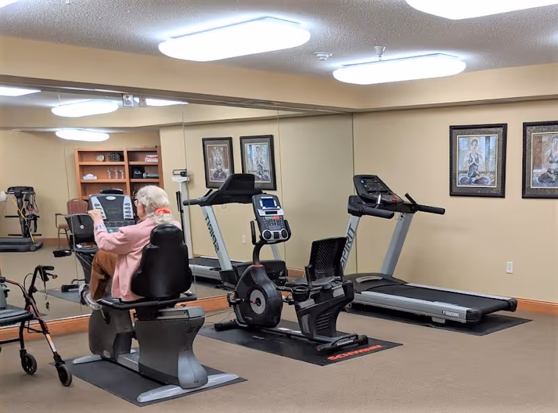 An elderly woman using a recumbent exercise bike in a fitness room with a treadmill and another exercise bike. The room has beige walls, a large mirror on one side, framed artwork on the walls, and fluorescent ceiling lights.