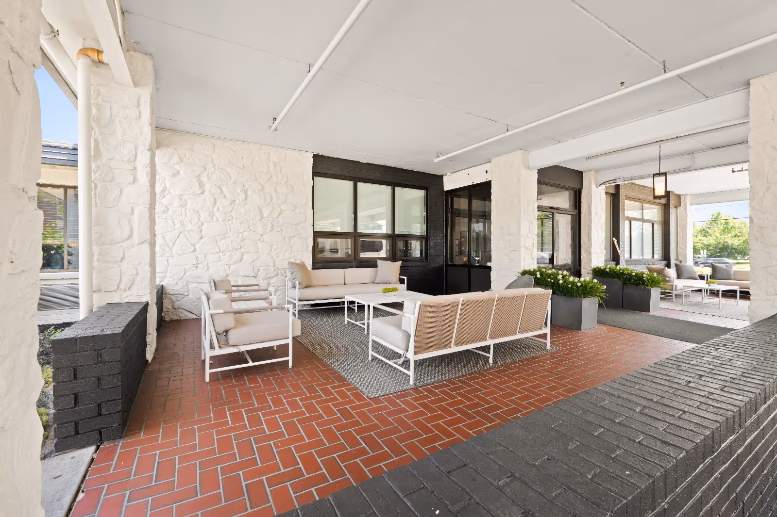 Covered outdoor seating area with cushioned sofas and chairs arranged around coffee tables on a red brick floor. The space is framed by white stone pillars and walls, with large windows and planters with green plants adding to the decor.