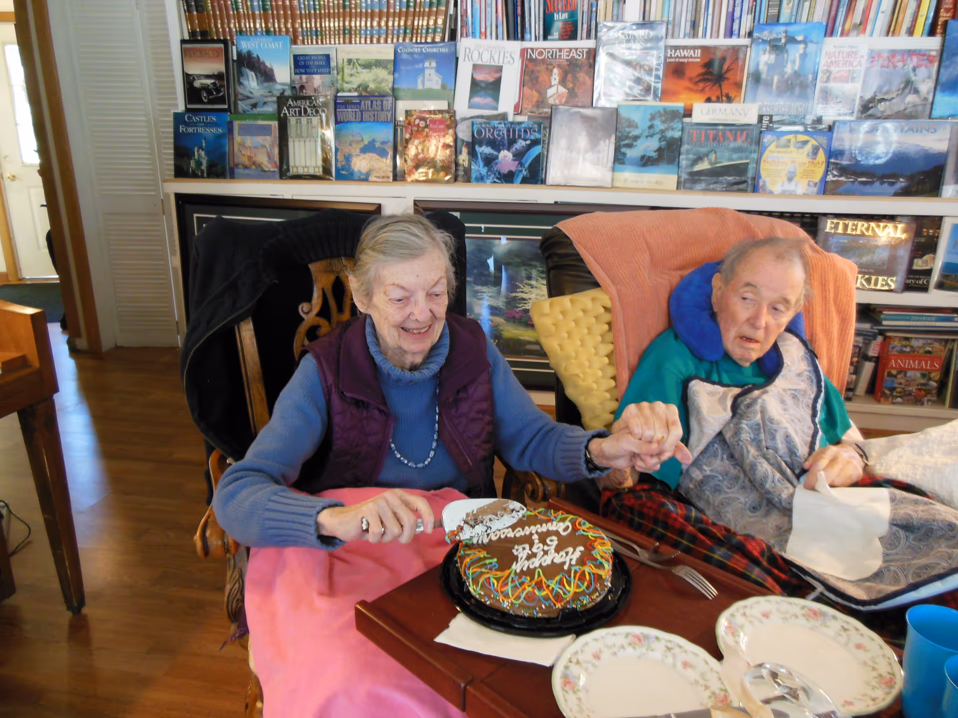 Two elderly residents holding hands while one cuts a decorated birthday cake at a table in a book-lined common room.