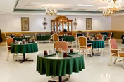 A spacious dining room with multiple round tables covered with green tablecloths, each set with cups and napkins. The room features beige upholstered chairs, chandeliers hanging from the ceiling, and a wooden cabinet against the far wall with decorative wall art on either side.