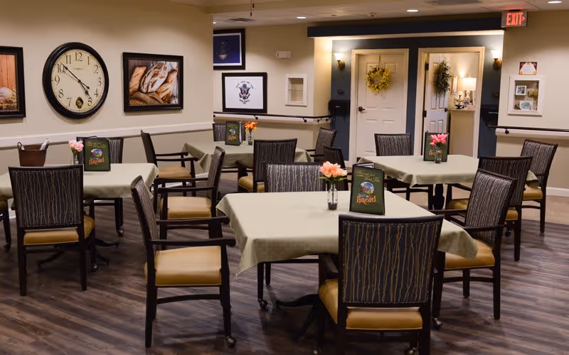 A dining room with several tables covered with light green tablecloths, each table having a small vase with pink flowers and a menu card labeled 'Bread'. The room has wooden chairs with cushioned seats, a large wall clock, framed pictures, and a door decorated with a wreath. The floor is dark wood, and the walls are light-colored with some dark trim.