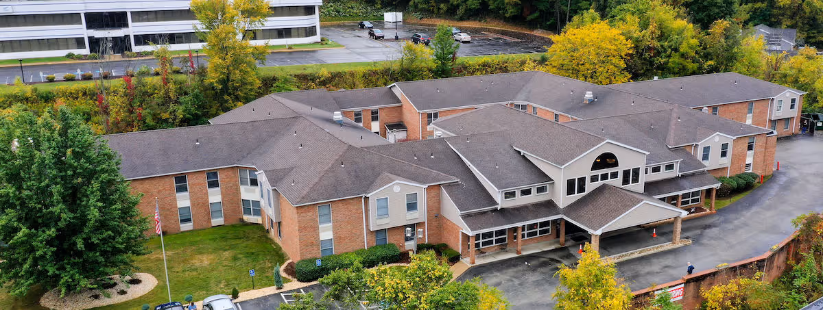 Aerial view of a large brick senior living facility with multiple connected wings and a covered entrance. The building is surrounded by trees with autumn foliage and a parking lot is visible in the background.