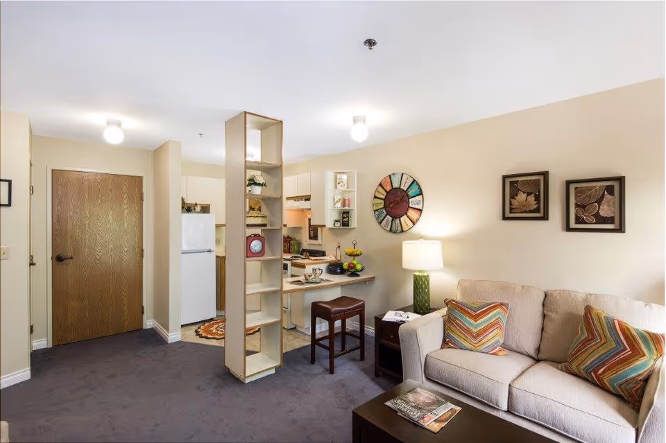 Interior view of a cozy living area in a retirement and assisted living facility. The room features a beige sofa with colorful chevron-patterned pillows, a wooden coffee table with magazines, a side table with a green lamp, and wall art including a colorful circular piece and framed leaf prints. A small kitchen area with white cabinets, a refrigerator, and a stove is visible in the background, partially separated by a narrow open shelving unit. The floor is carpeted in a dark color, and the walls are painted a light beige.