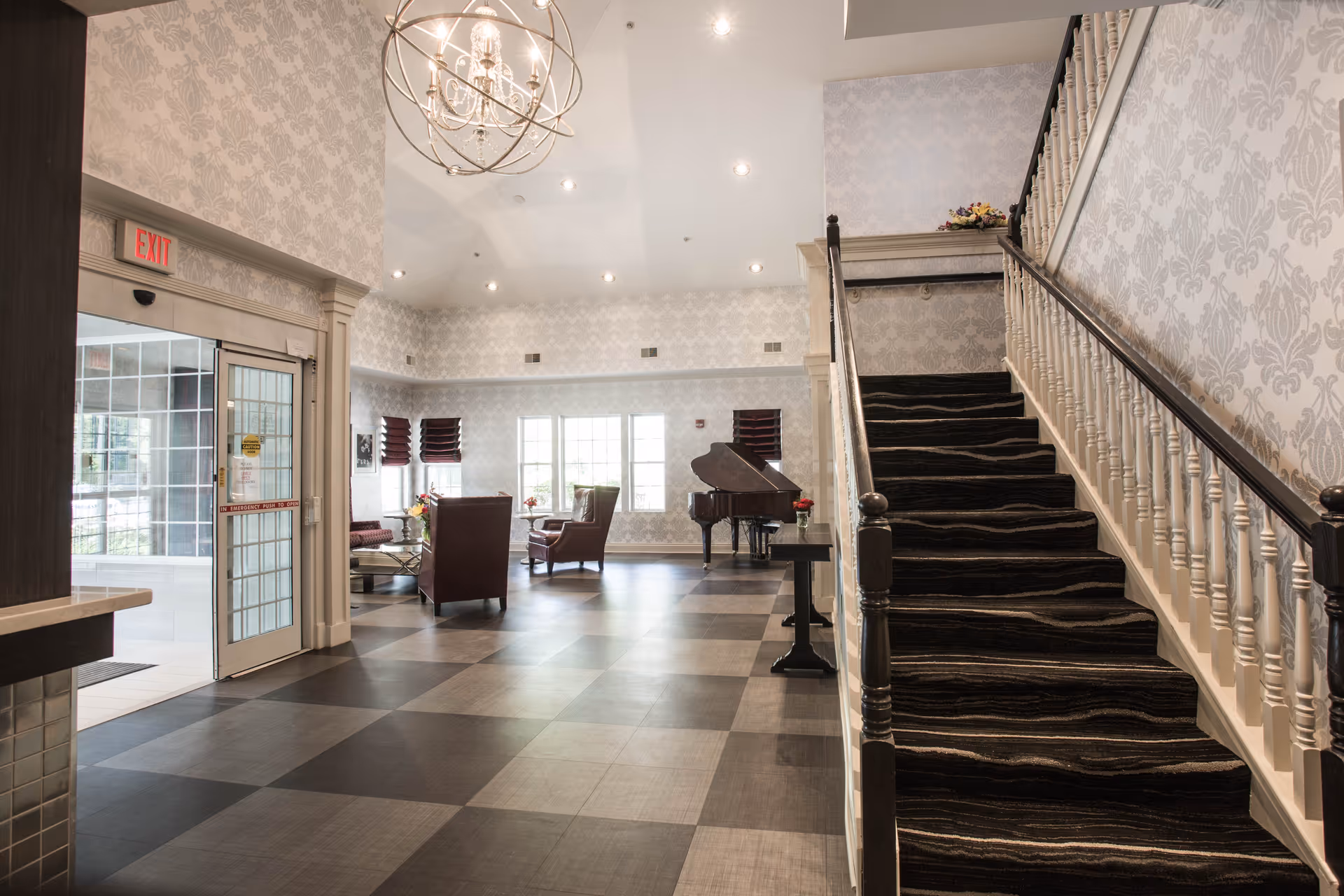 Spacious assisted-living lobby with a carpeted staircase on the right, seating area and grand piano in the center, and glass entrance doors on the left.