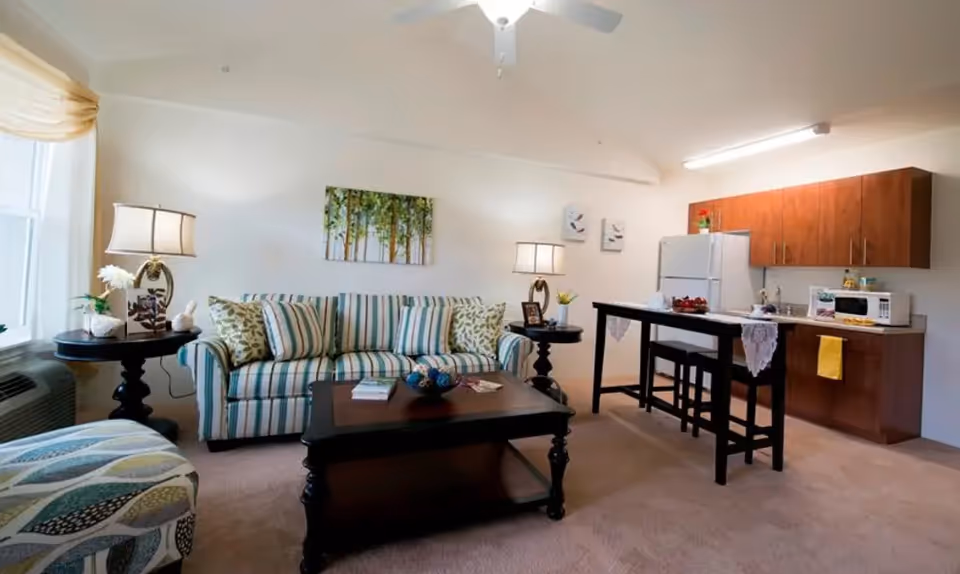 A bright living area in an assisted living facility featuring a striped sofa with patterned pillows, a dark wooden coffee table, two side tables with lamps, and a kitchen area with wooden cabinets, a white refrigerator, microwave, and a high table with two stools.