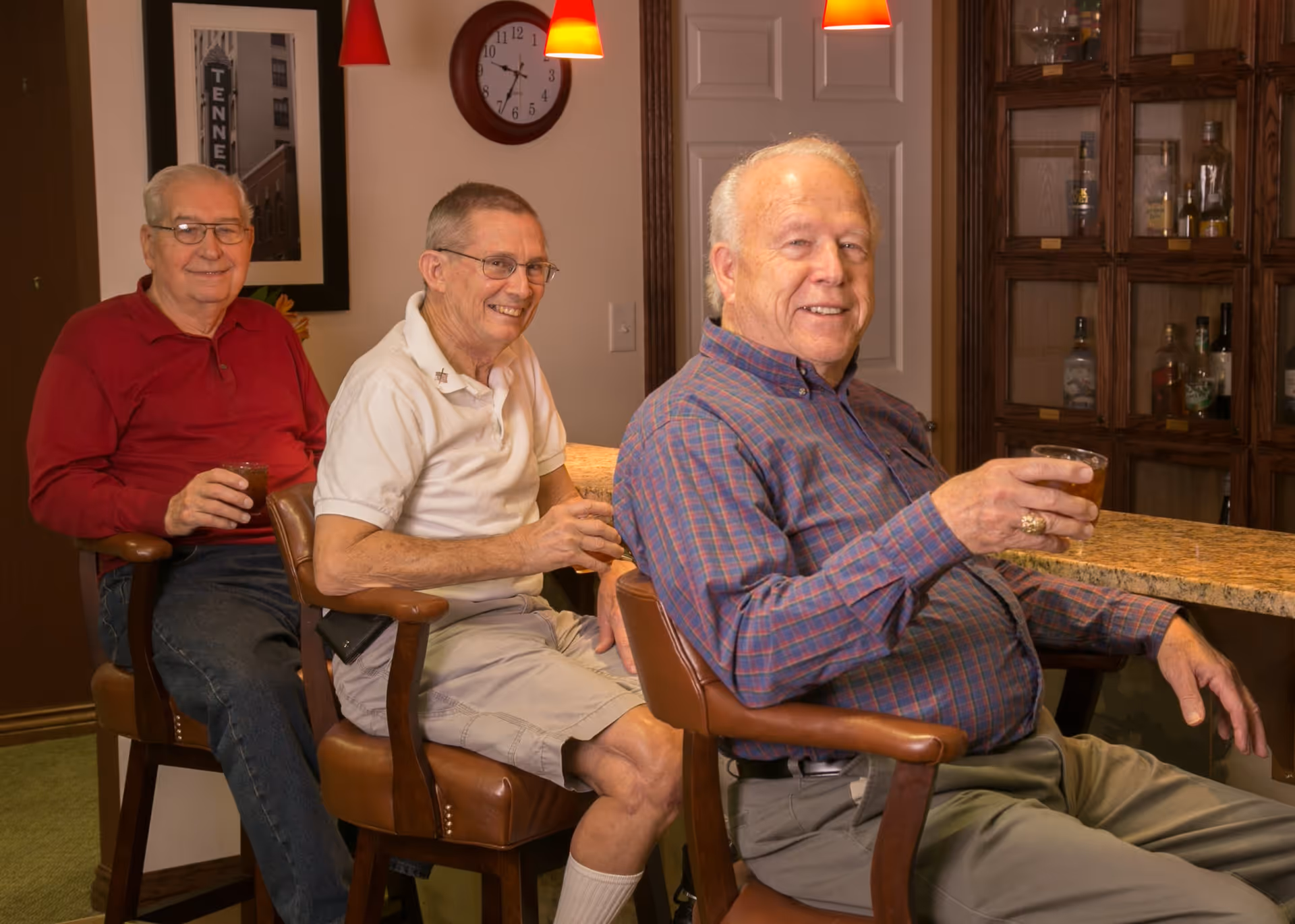 Three elderly men sitting on bar stools at a counter in a cozy indoor setting, each holding a drink and smiling. Behind them is a wooden cabinet with bottles and glassware, a clock on the wall, and red pendant lights hanging from the ceiling.