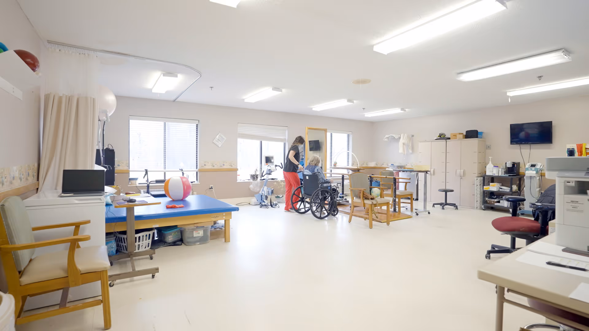 A bright and spacious therapy room in a healthcare facility with large windows letting in natural light. The room contains various equipment including a therapy table with a beach ball on it, chairs, a wheelchair, and exercise machines. A caregiver is assisting an elderly person in a wheelchair. The walls are light-colored with a decorative border, and there are cabinets and a TV mounted on the wall.