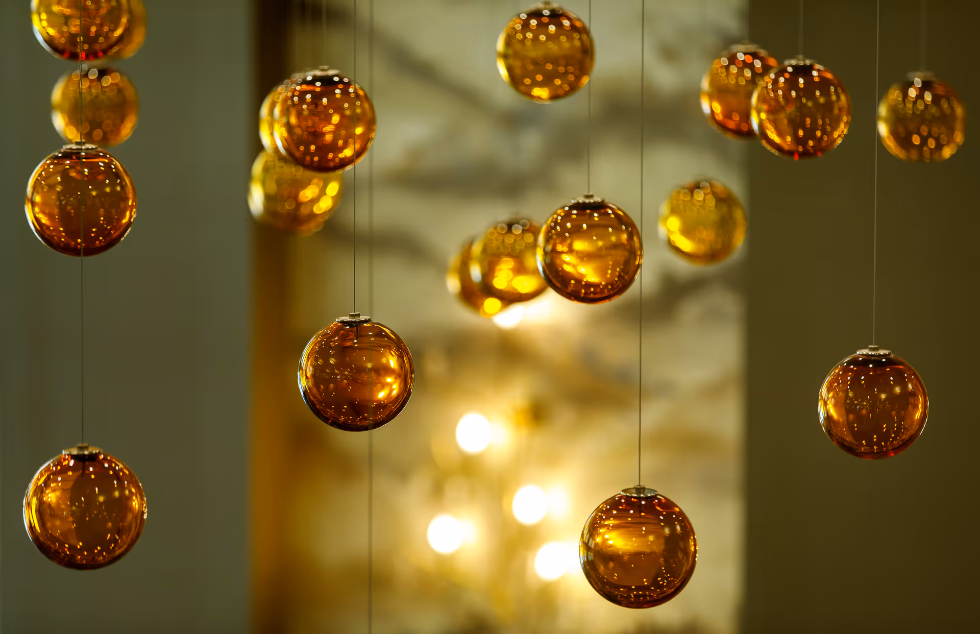 Close-up view of multiple amber-colored glass spheres hanging from thin strings, with a softly lit, blurred background featuring warm light spots.