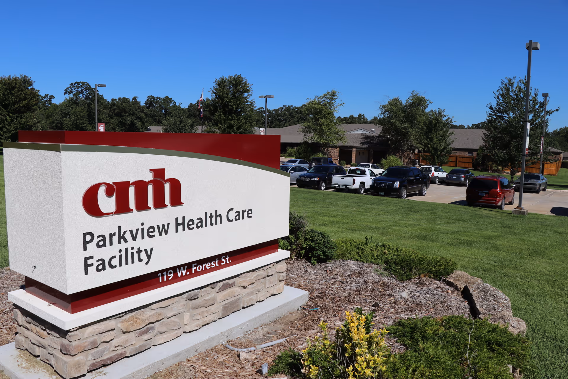 A stone monument sign reading "Parkview Health Care Facility" in front of a parking lot and low-rise building under a clear blue sky.