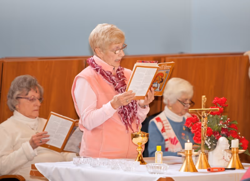 Three elderly women participating in a religious service inside a room. One woman in a pink vest and scarf is standing and reading from a booklet, while the other two women are seated and also reading. A table in front of them is covered with a white cloth and holds religious items including a gold chalice, candles, a crucifix, flowers, and a small bottle of hand sanitizer.