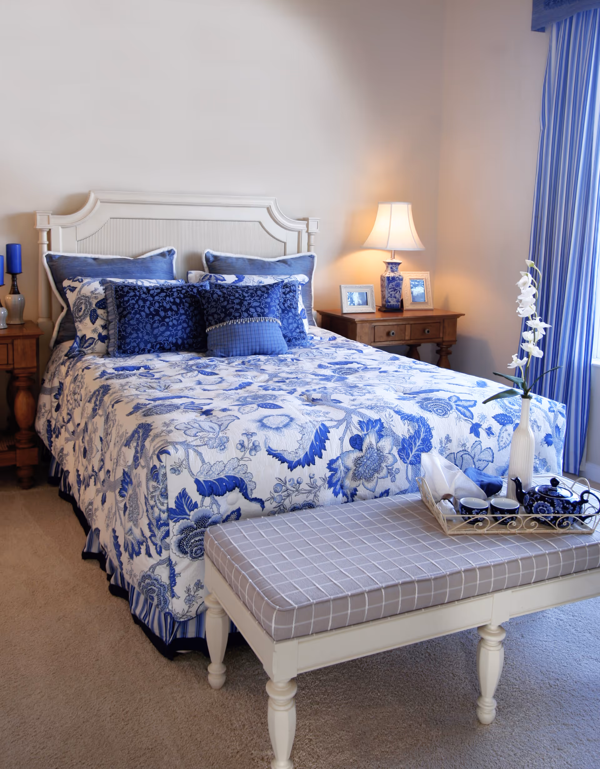 A neatly made bed with a white headboard and blue floral bedding in a bedroom. The bed has multiple blue and white pillows. There is a wooden nightstand with a lamp and framed photos on the right side. A cushioned bench with a gray checkered pattern is at the foot of the bed, holding a tray with a teapot, cups, and a white vase with flowers. Blue curtains hang by the window on the right.