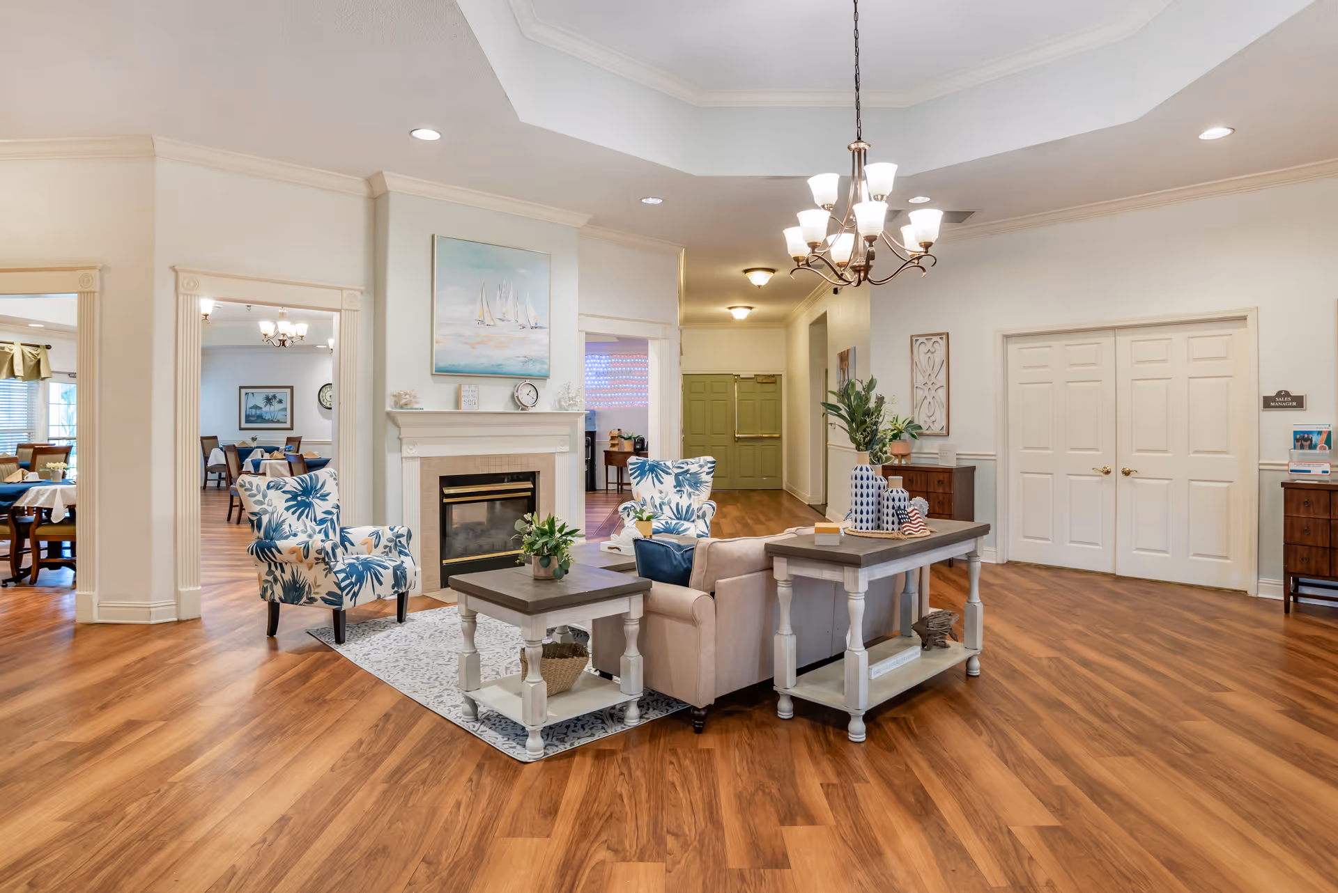 Bright communal living room with sofas, patterned armchairs, a fireplace, and a central chandelier.
