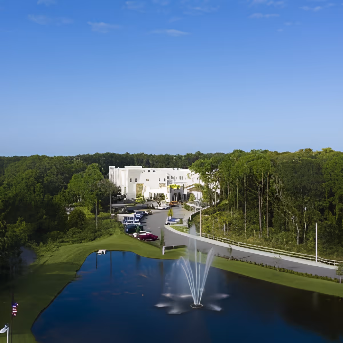 Aerial view of Discovery Village At Westchase facility surrounded by dense green trees, with a clear blue sky above. In the foreground, there is a pond with a water fountain spraying water upwards. A driveway leads to the white building complex with several parked cars along the side.