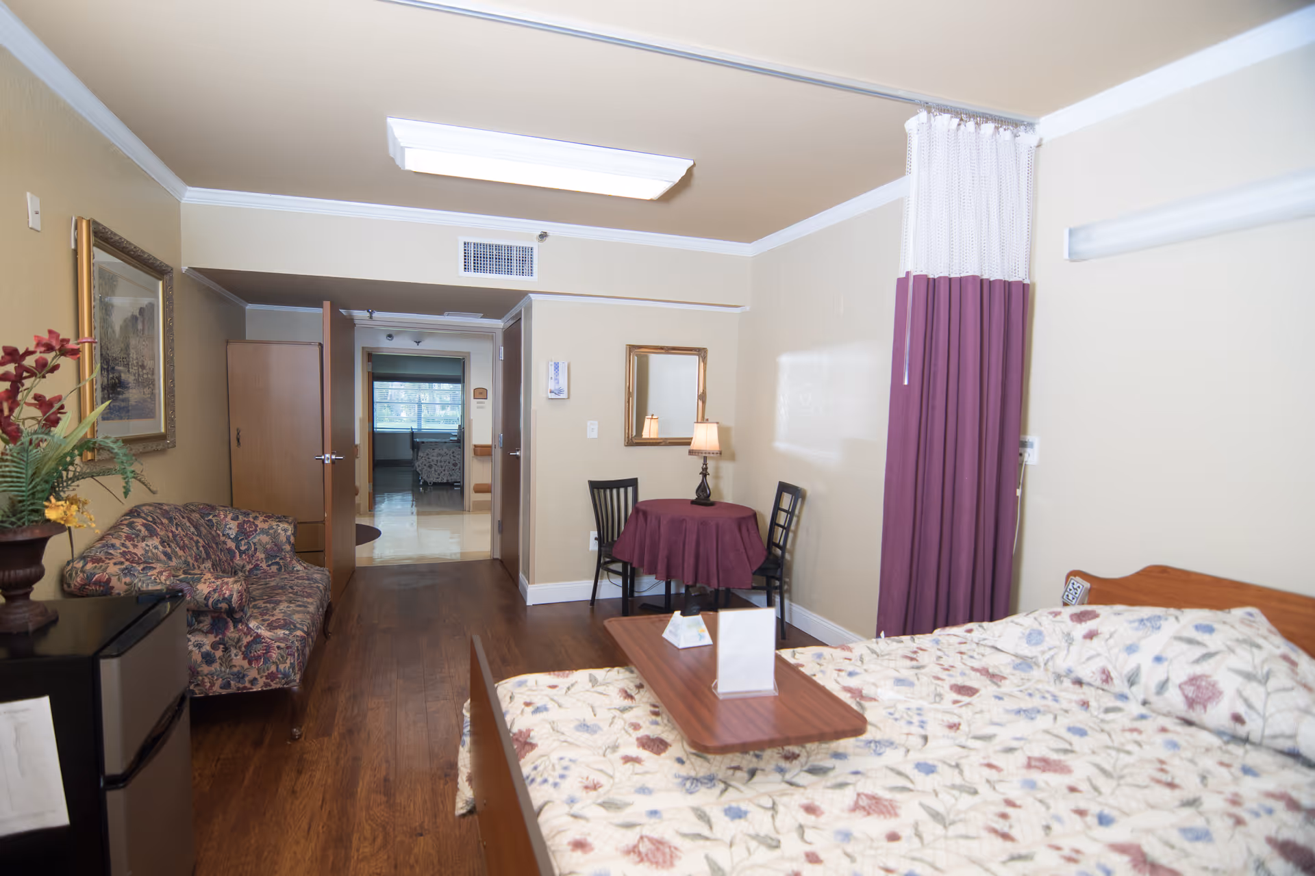 A senior living facility bedroom with a floral-patterned bedspread on a bed, a small table with two chairs and a lamp, a floral couch, a mini refrigerator, and a curtain divider. The room has wooden flooring and beige walls with a framed picture and a mirror.
