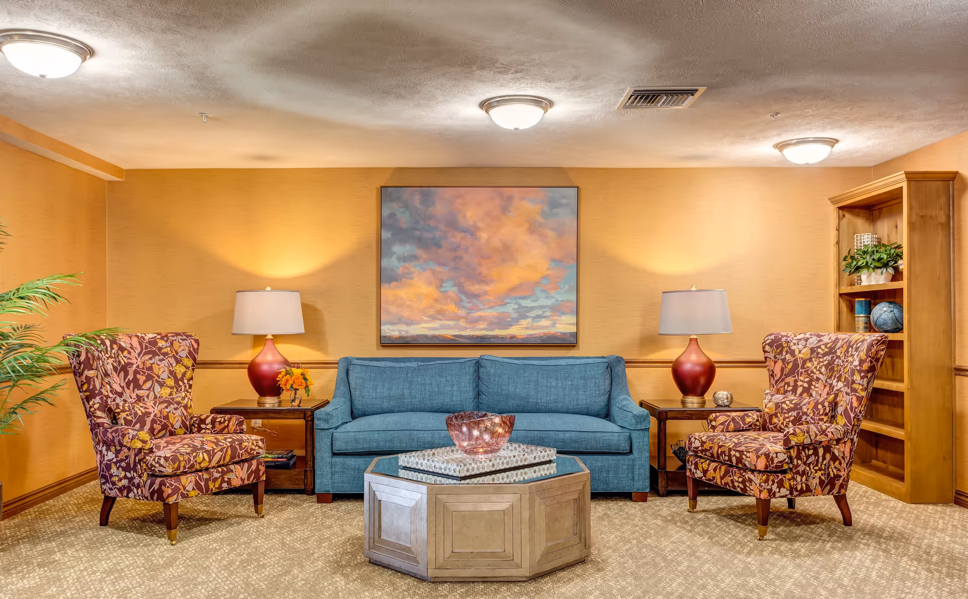 A cozy living room area with a blue sofa centered between two floral-patterned armchairs. Each armchair is next to a wooden side table with a red lamp and decorative items. A large colorful painting of a cloudy sky hangs on the wall behind the sofa. There is a wooden bookshelf with plants and decorative objects on the right side. The room has warm yellow walls and a beige carpeted floor.