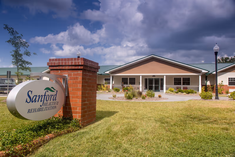 Front entrance of Sanford Health & Rehabilitation building with a sign on a brick post, lawn, and cloudy sky.