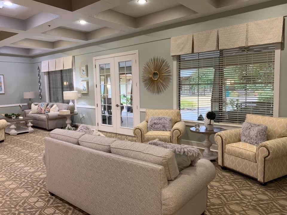 A cozy living room area in an assisted living facility featuring beige sofas and armchairs with patterned cushions, a patterned carpet, two side tables with lamps and decorative items, large windows with blinds, and French doors leading to another room.