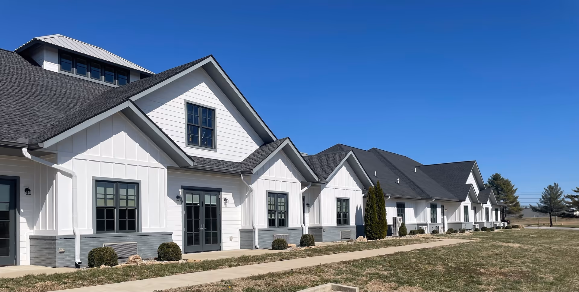 Exterior view of a single-story senior living facility building with white siding, gray trim, multiple windows, and a dark shingled roof under a clear blue sky. There is a concrete walkway and some small bushes in front of the building.