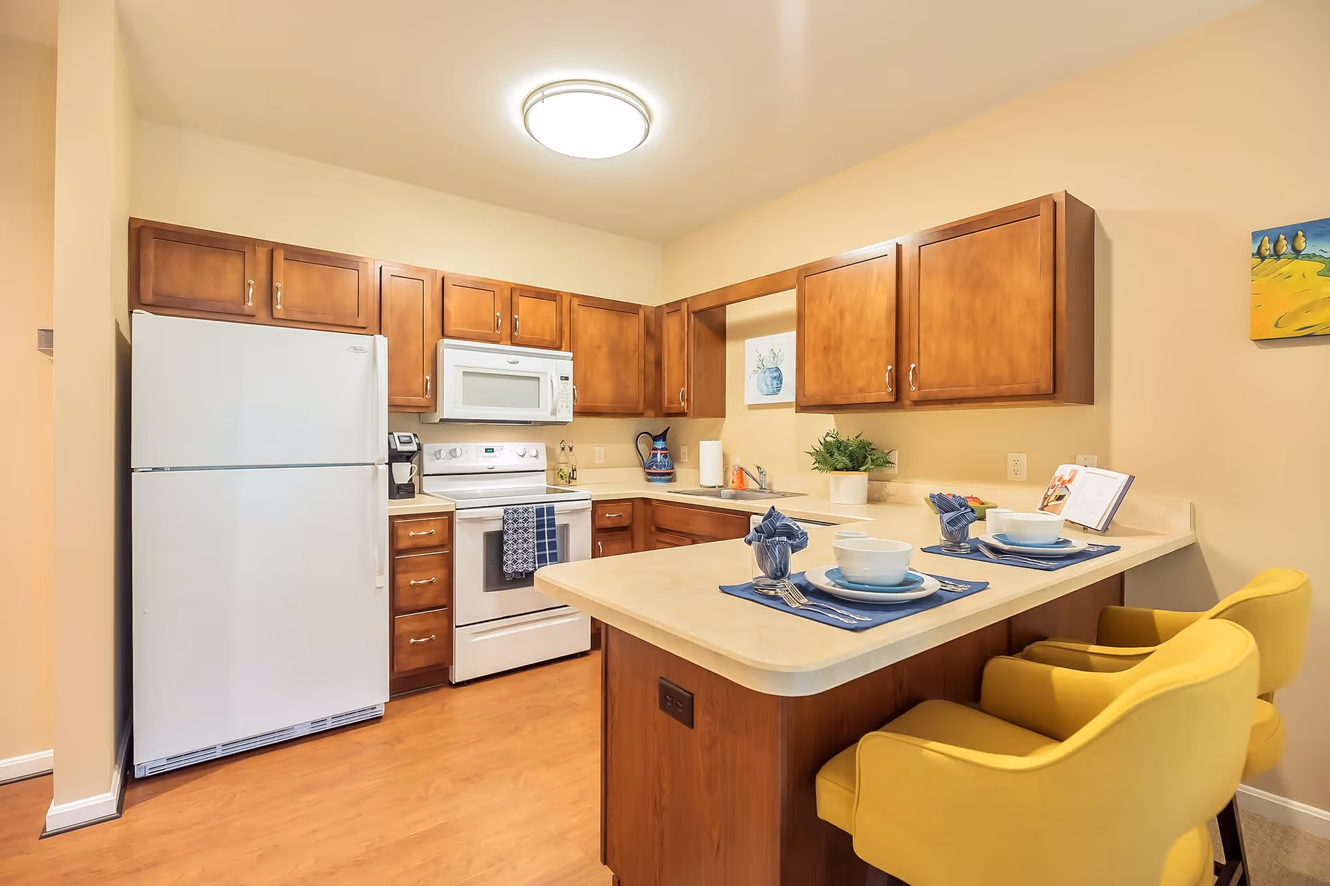 A bright kitchen with wooden cabinets, a white refrigerator, stove, and microwave. There is a countertop with two place settings, each with a bowl, plate, utensils, and a folded napkin. Two yellow chairs are positioned at the counter. The walls are light beige, and there is a small plant and a painting on the wall.