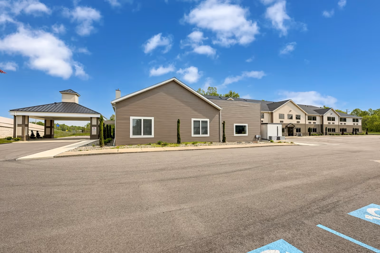 Exterior view of a senior living facility building with a large parking lot in front, a covered entrance on the left, and a clear blue sky with some clouds above.