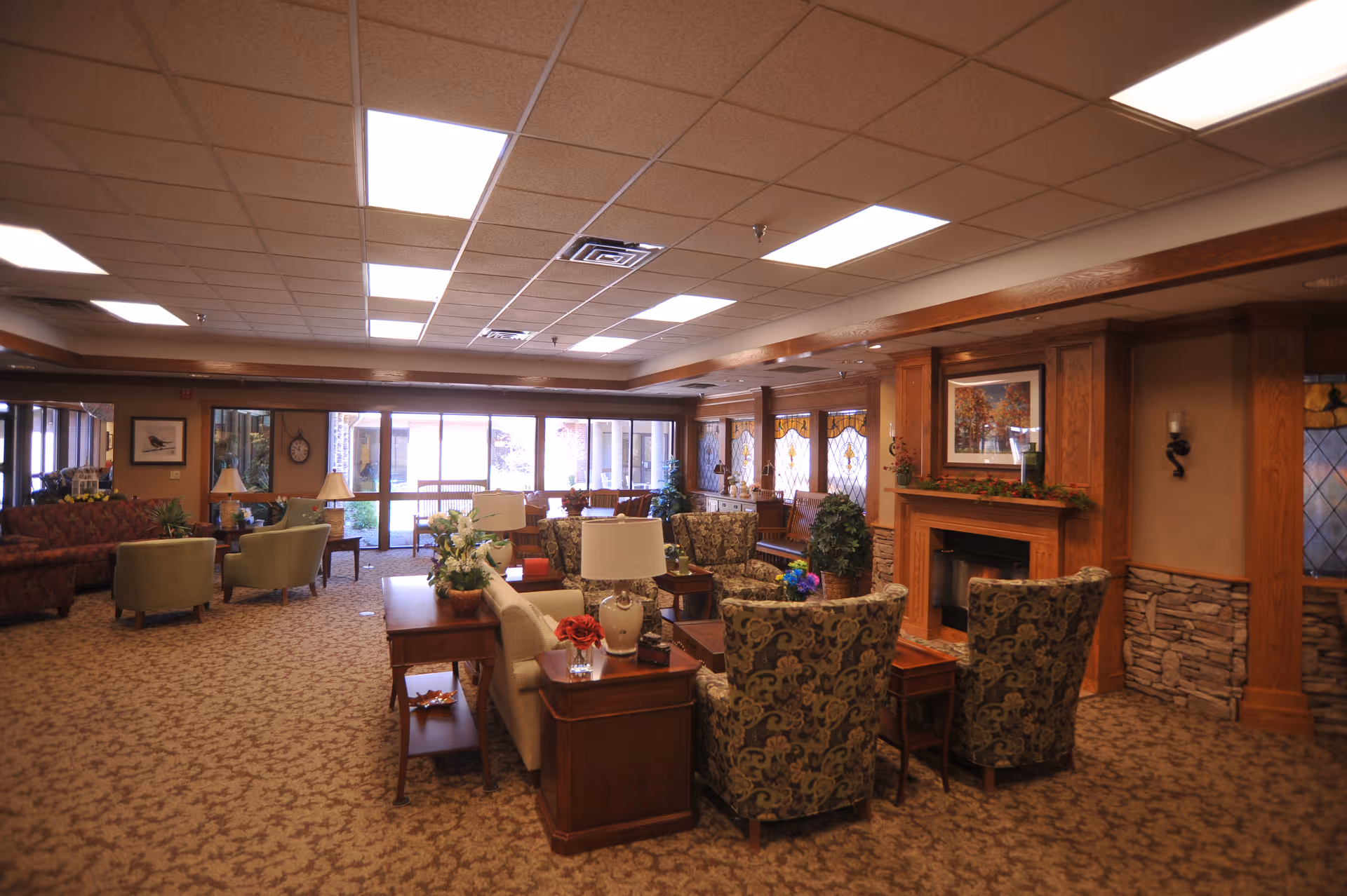 A spacious and warmly decorated living room area in an assisted living facility, featuring multiple upholstered chairs and sofas arranged around wooden tables and a fireplace. The room has carpeted floors, a drop ceiling with fluorescent lighting, large windows with stained glass accents, and various plants and framed artwork on the walls.