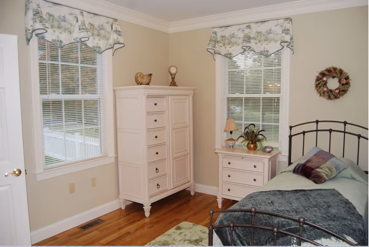 A cozy bedroom with two windows covered by patterned valances. The room features a white wooden dresser and a matching nightstand with a small lamp and decorative items. There is a single bed with a metal frame, a green blanket, and a multicolored pillow. The floor is wooden, and the walls are painted beige.