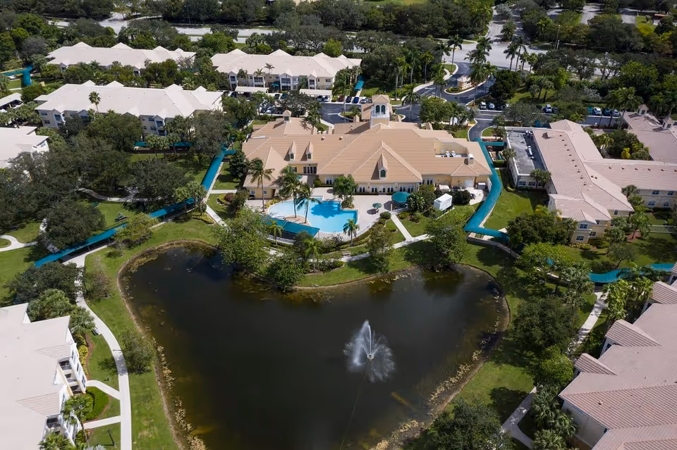 Aerial view of a senior living community with a central clubhouse and pool, surrounding residential buildings, walkways and a pond with a fountain.