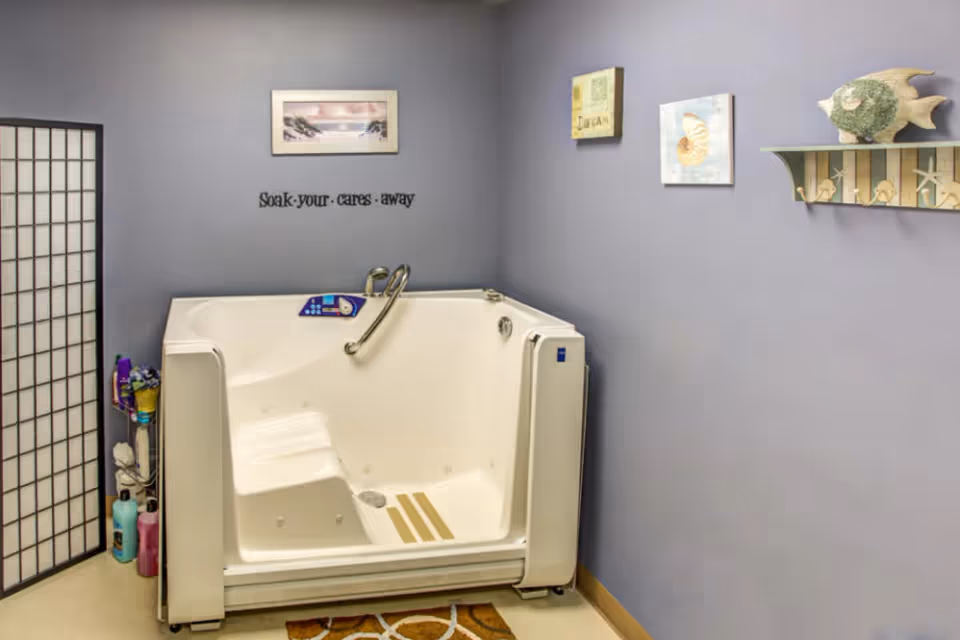A walk-in bathtub with a built-in seat and handrail in a room with light purple walls. The wall above the tub has a framed picture, decorative signs, and a shelf with sea-themed decorations. There is a folding screen on the left side and various bath products on the floor next to the tub.