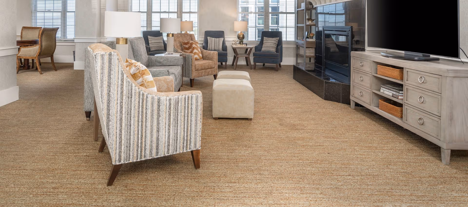 Bright communal living room with sofas, armchairs, ottomans, a TV console, and large windows.
