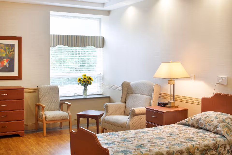 Sunlit bedroom with a single bed, nightstand and lamp, two chairs, a small table and a window with a vase of sunflowers.