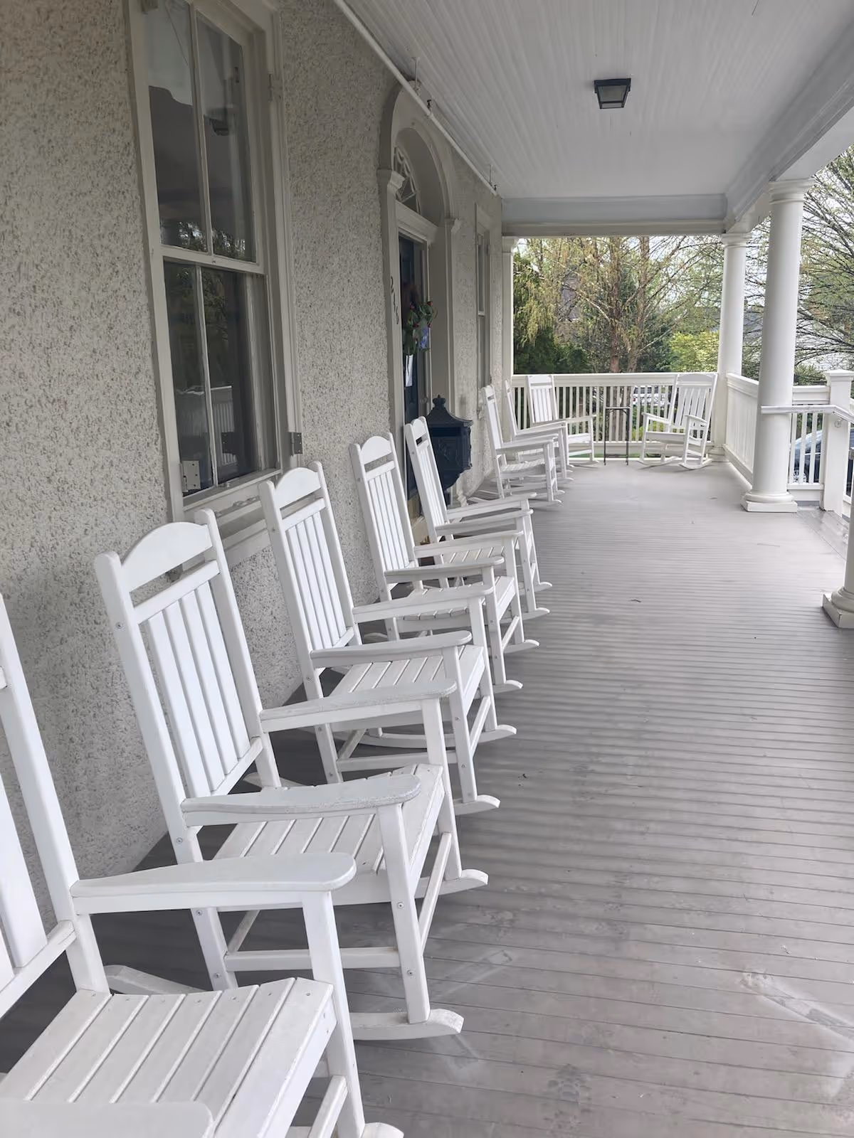 A long covered porch with a row of white wooden rocking chairs lined up along the wall of a building. The porch has a light gray floor and white railing with columns, overlooking a green outdoor area with trees.