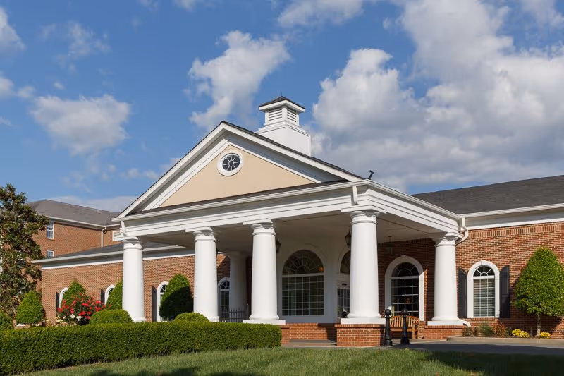 Front exterior view of a brick building with white columns supporting a covered entrance, surrounded by neatly trimmed bushes and trees under a partly cloudy sky.
