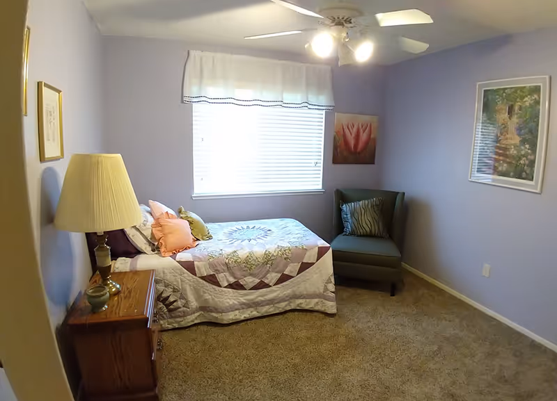 Small bedroom with a single bed covered by a quilt, a wooden nightstand with a lamp, an upholstered chair, a window with blinds, and a ceiling fan.
