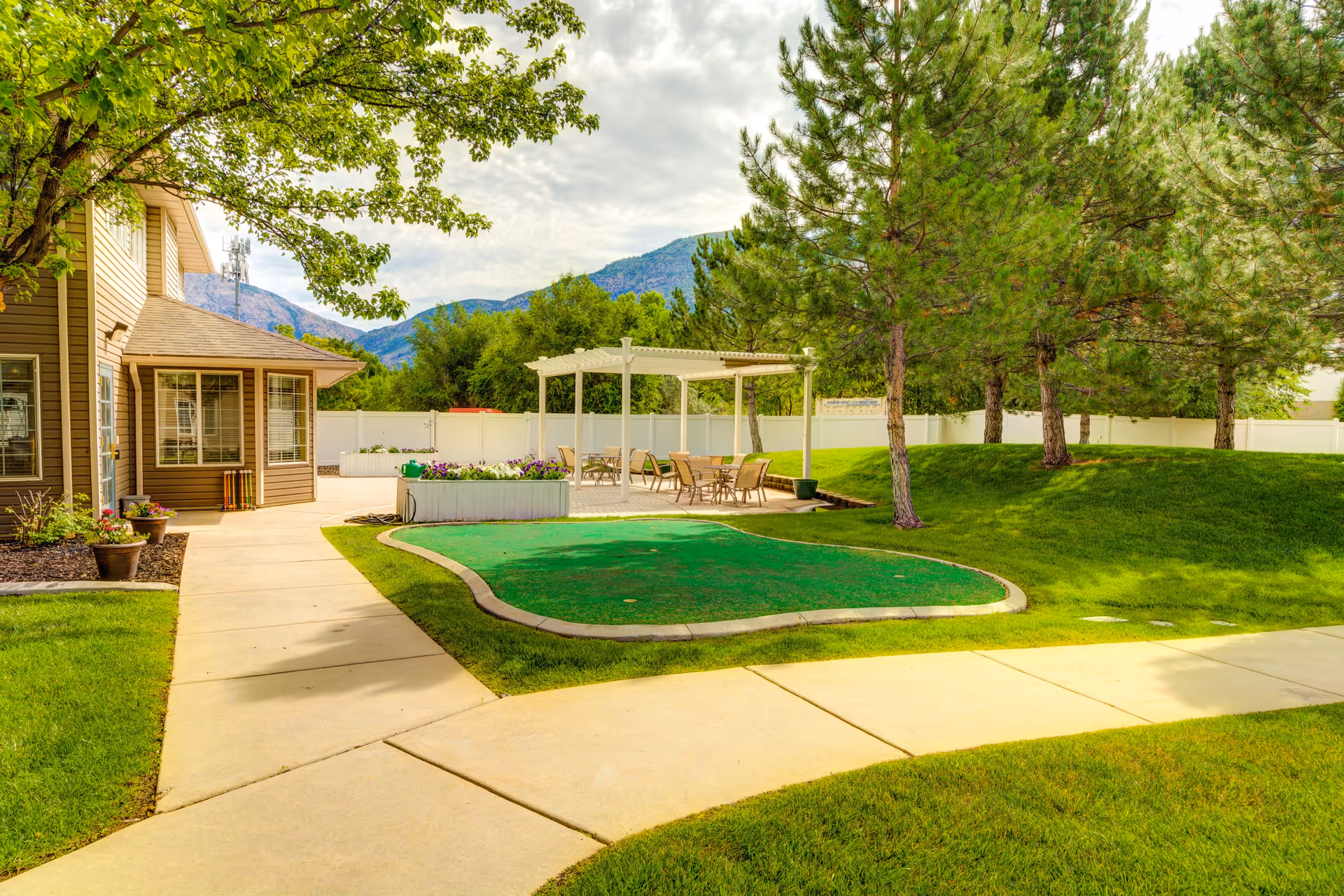 Outdoor area of a senior living facility with a paved walkway, green grass, trees, a white pergola with tables and chairs underneath, and a building on the left side. Mountains and a partly cloudy sky are visible in the background.