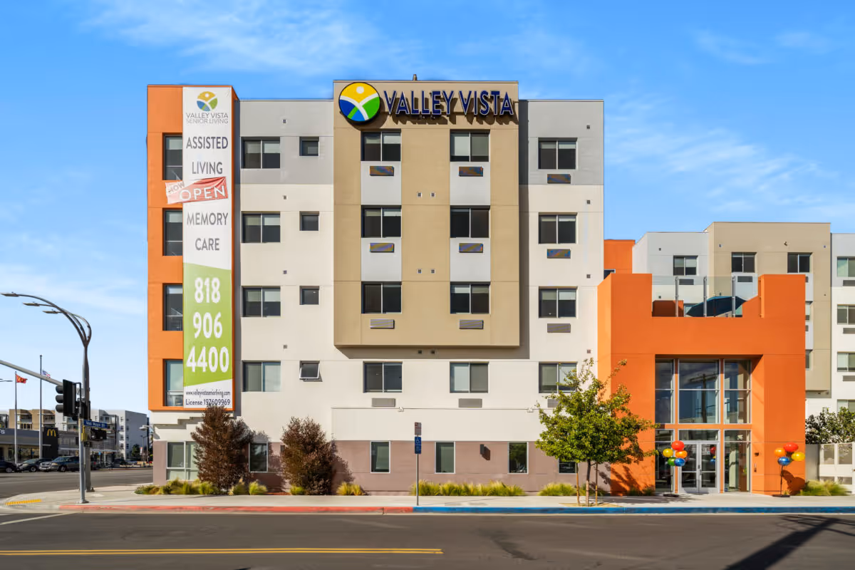 Front exterior view of the Valley Vista Senior Living building, a multi-story structure with a beige, white, and orange facade. The building has multiple windows and a large sign with the Valley Vista logo and name at the top center. A vertical banner on the left side advertises assisted living and memory care services with a phone number. There are trees and balloons near the entrance, and a clear blue sky in the background.