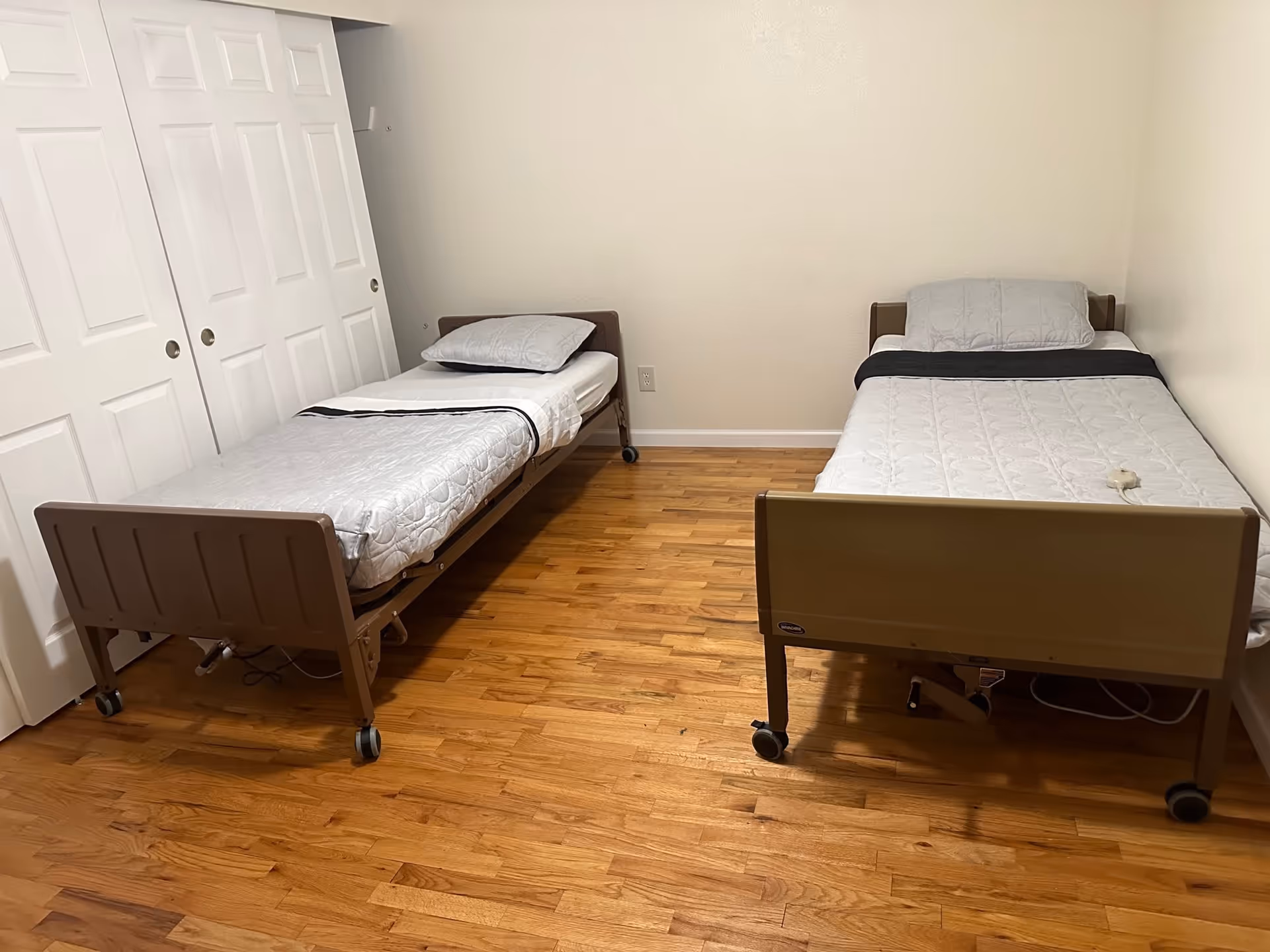 A simple bedroom with two single hospital-style beds with metal frames and wheels, each made with white bedding and a gray pillow. The room has wooden flooring, beige walls, and white sliding closet doors on the left side.
