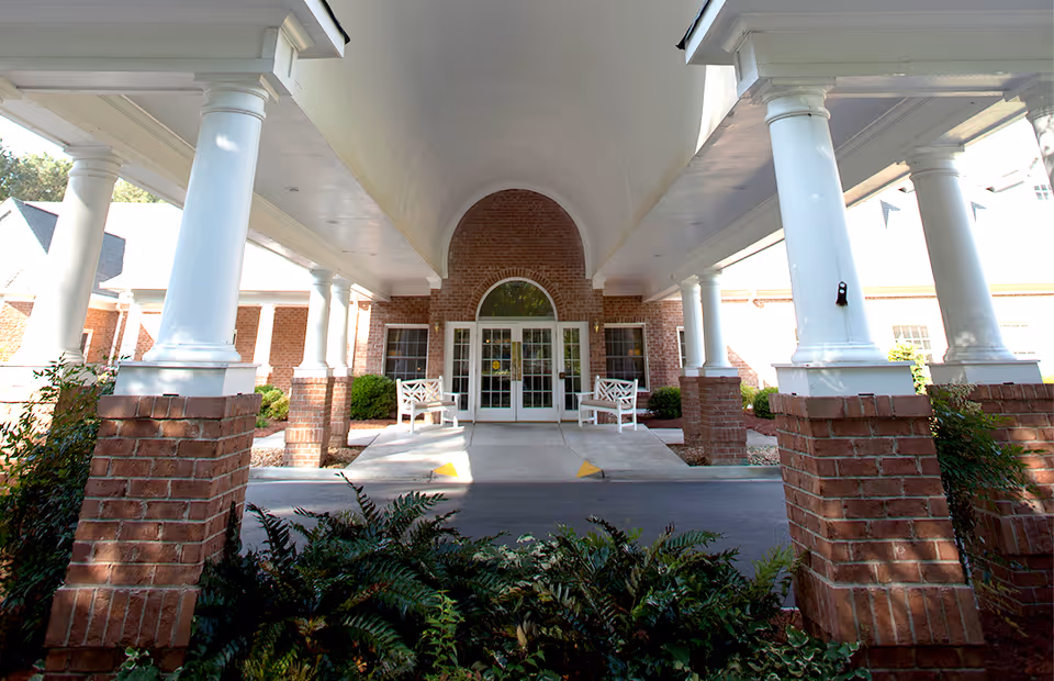Entrance of a senior living facility with a covered driveway supported by white columns on brick bases. Two white benches are placed near the glass double doors, with greenery and shrubs around the area.