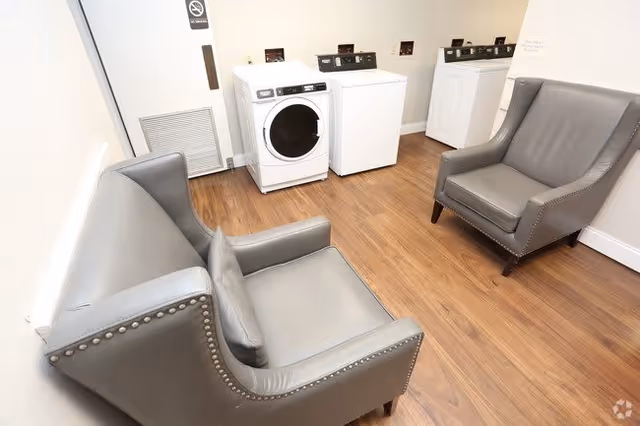 A small laundry room with two white washing machines and one white dryer against the wall. The room has wood flooring and white walls. There are two gray leather armchairs with nailhead trim positioned facing each other in the foreground.