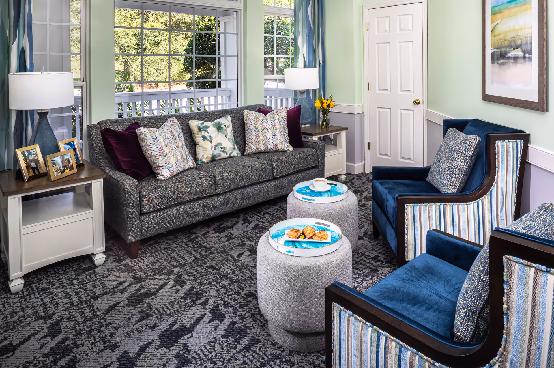 Cozy living room with a gray sofa, two blue armchairs, round ottomans with trays, side tables and a large window overlooking greenery.