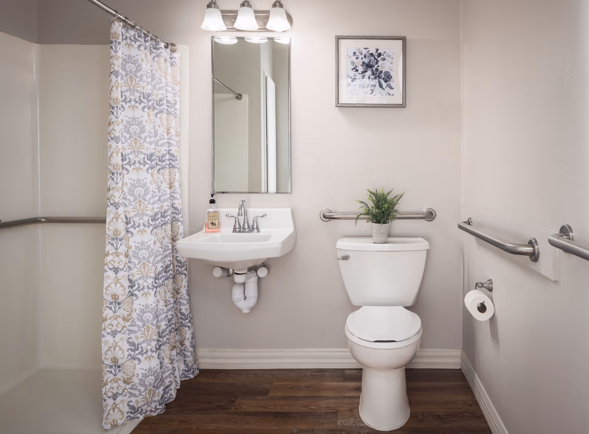 A clean and accessible bathroom featuring a white toilet with a small potted plant on top, a wall-mounted sink with a soap dispenser, a mirror above the sink, a shower area with a patterned curtain, and multiple grab bars for support. The floor is wood-style, and a framed floral artwork hangs on the wall above the toilet.