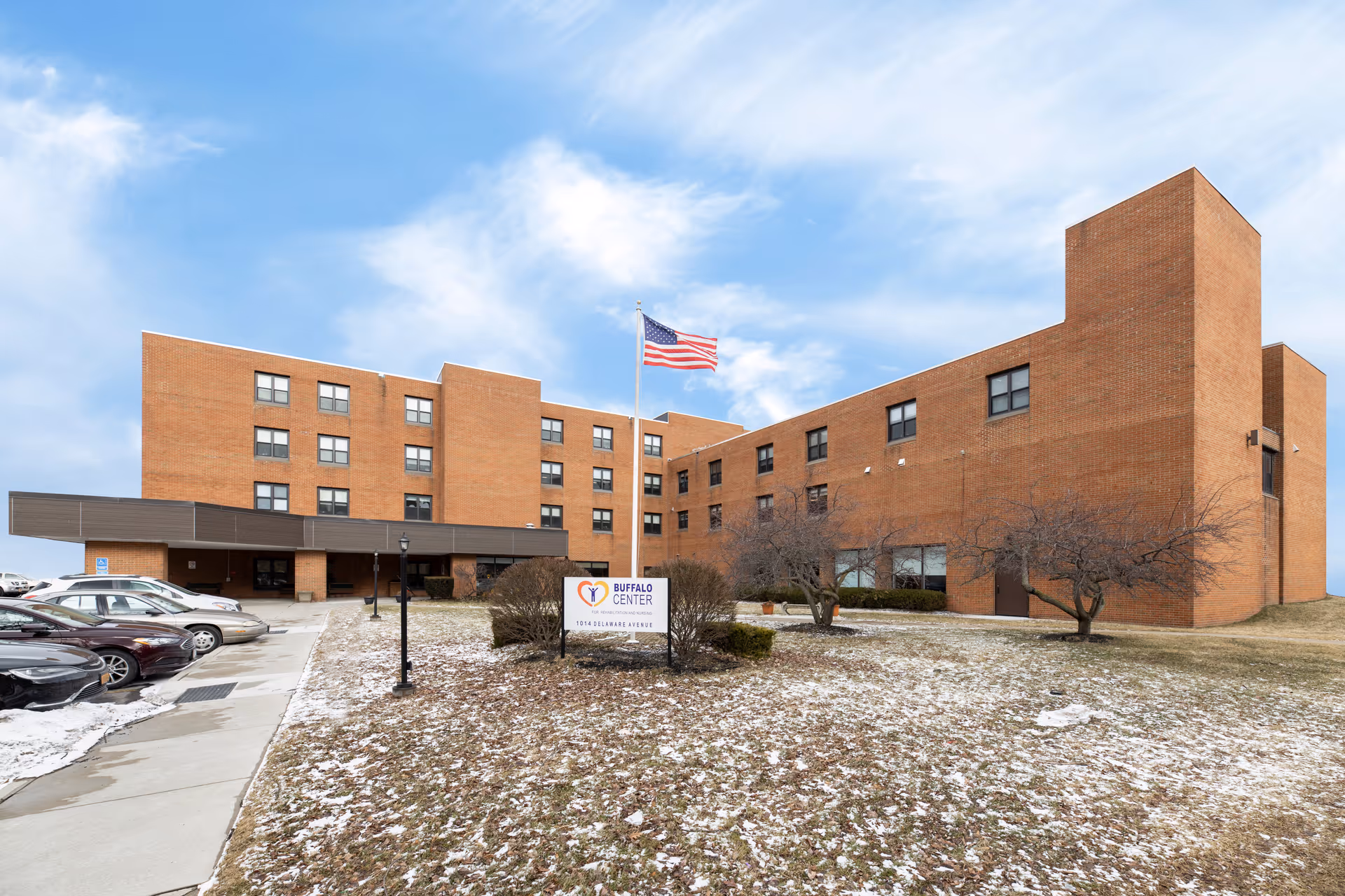 Exterior view of a multi-story brick building with an American flag on a flagpole in front. There is a sign that reads 'Buffalo Center' near the entrance, and several parked cars are visible along the driveway. The ground has patches of snow and some leafless trees and bushes.