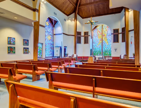 Interior view of a chapel with wooden pews arranged in rows facing a large stained glass window and a cross at the front. The ceiling is high with wooden beams, and additional stained glass windows and religious artwork are visible on the walls.