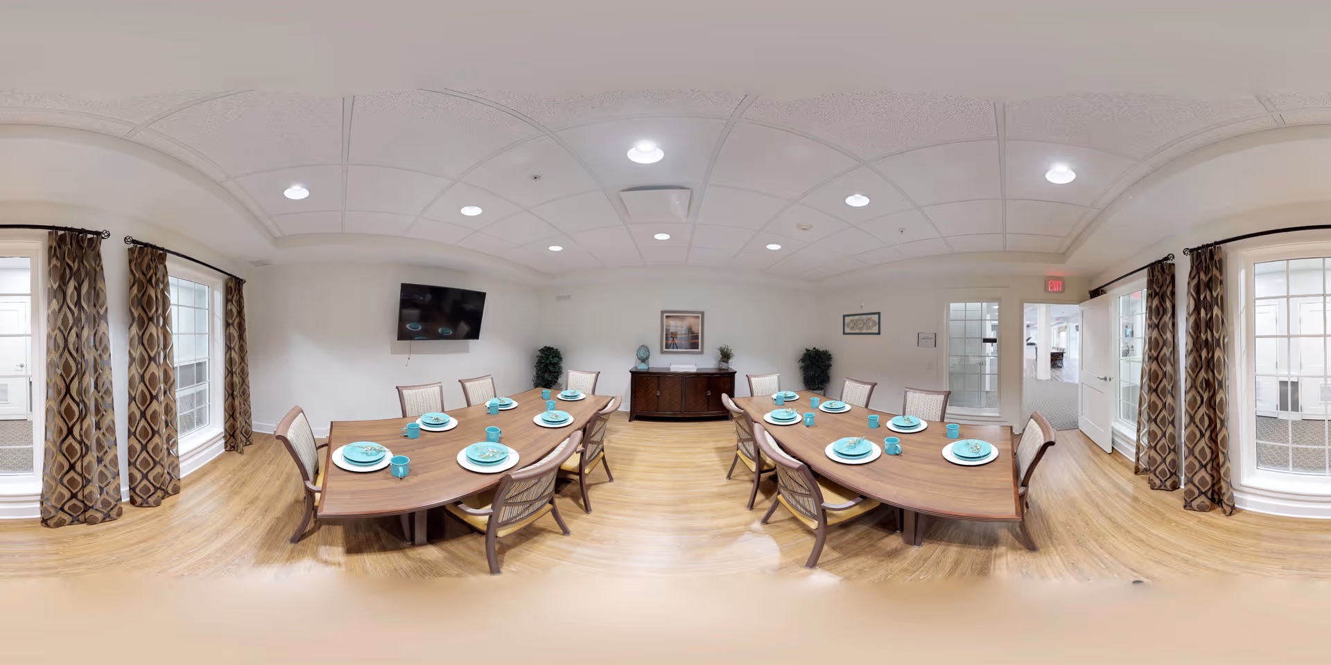 A bright dining room with two wooden tables arranged in a U-shape, each set with turquoise plates, bowls, and mugs. The room has wooden flooring, white walls, and large windows with patterned curtains allowing natural light to enter. A flat-screen TV is mounted on one wall, and there are decorative plants and framed artwork on the walls. The ceiling has recessed lighting, and there is a door leading to another room.