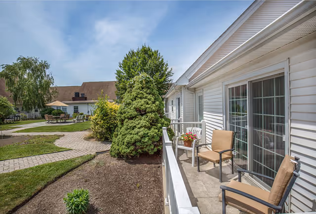 Outdoor patio area at a senior living facility with two cushioned chairs and a small table with a flower pot, adjacent to a white building with sliding glass doors. The patio overlooks a landscaped garden with a paved walkway, green bushes, trees, and outdoor seating with an umbrella in the background under a partly cloudy sky.
