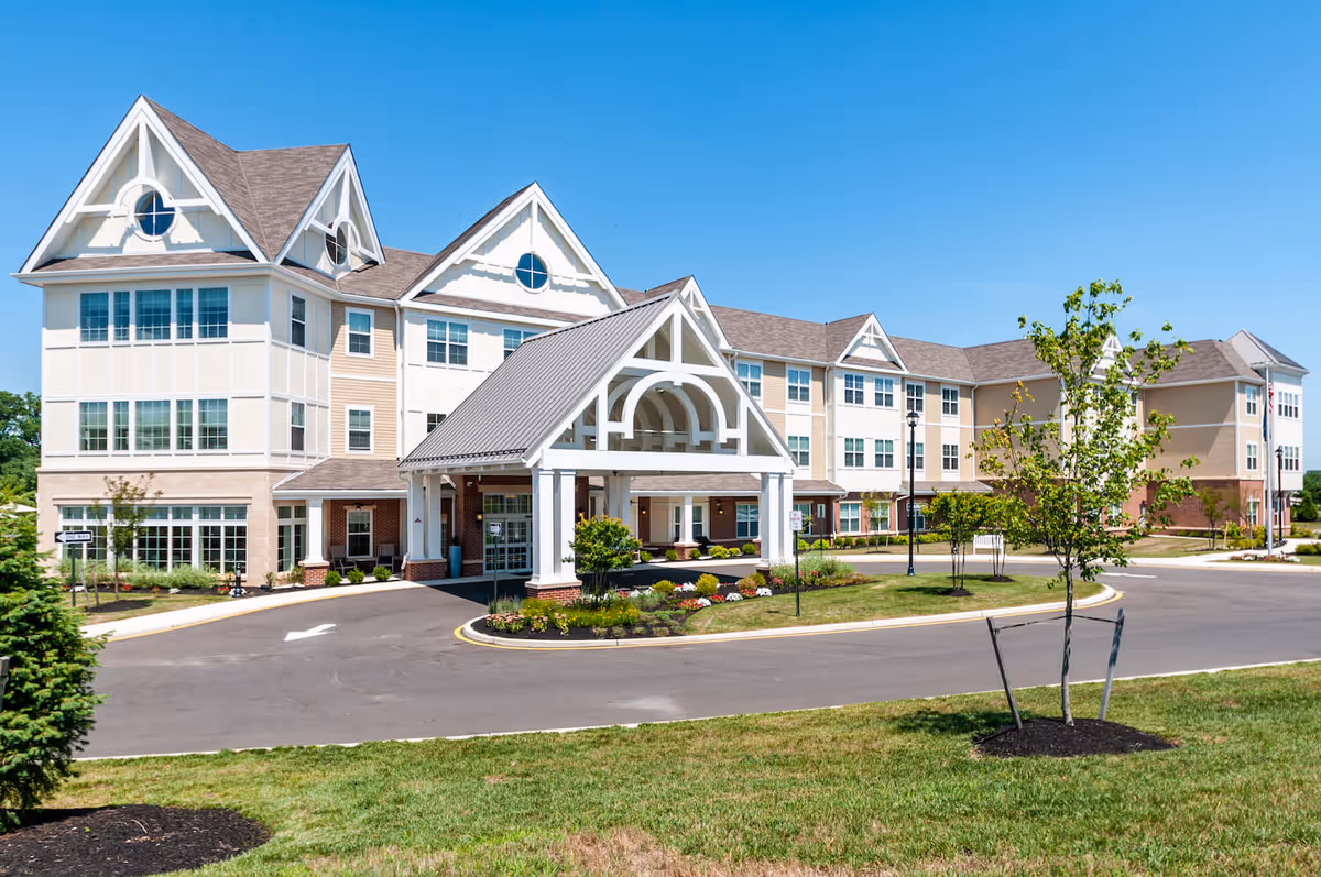 Exterior view of a large, multi-story senior living facility building with a covered entrance, manicured landscaping, and a clear blue sky.