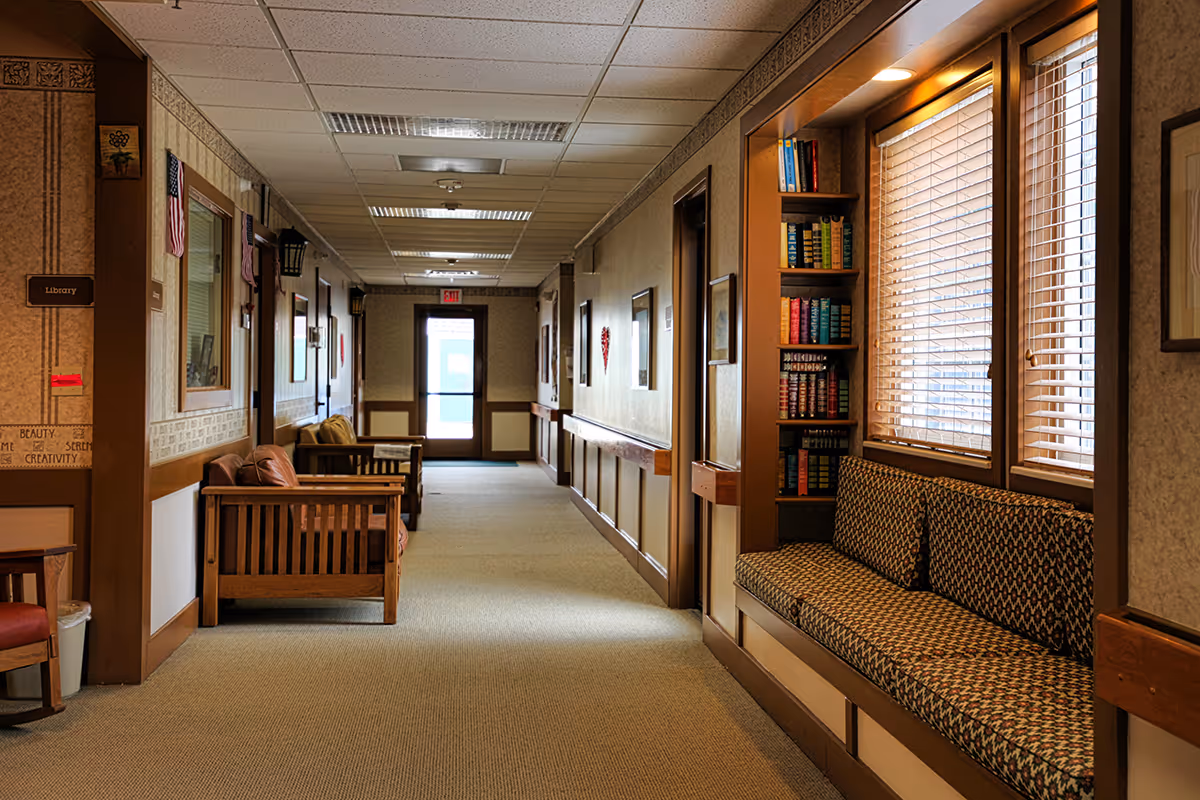 A carpeted interior hallway with wooden benches, a built-in bookcase, and windows with blinds in a senior living facility.