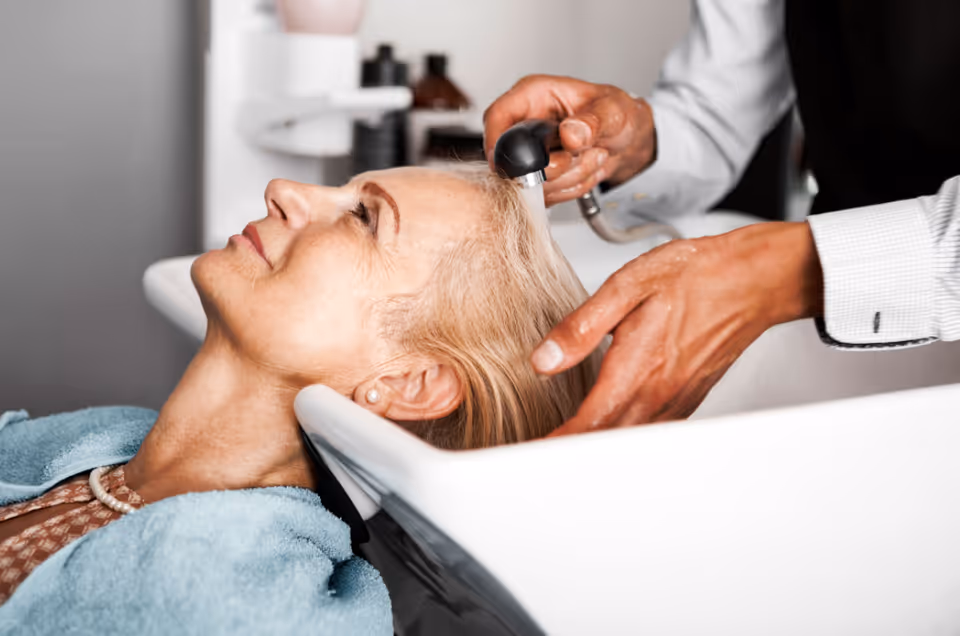 An elderly woman with blonde hair is reclining with her head in a salon sink while a person washes her hair using a handheld sprayer.