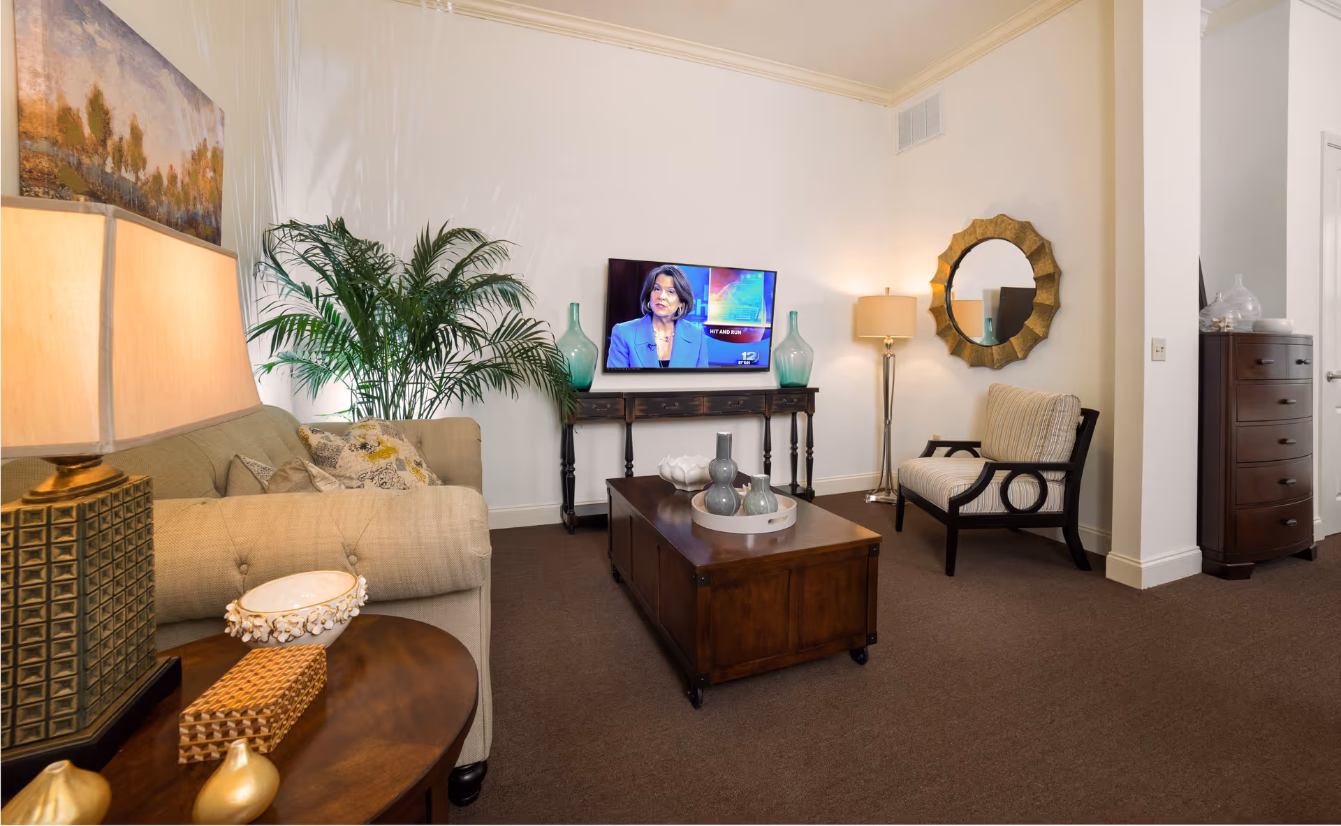 A cozy living room with a beige sofa, a wooden coffee table with decorative vases, a cushioned armchair, a flat-screen TV mounted on a wooden console table, a large green plant, a round decorative mirror on the wall, and two lamps providing warm lighting.