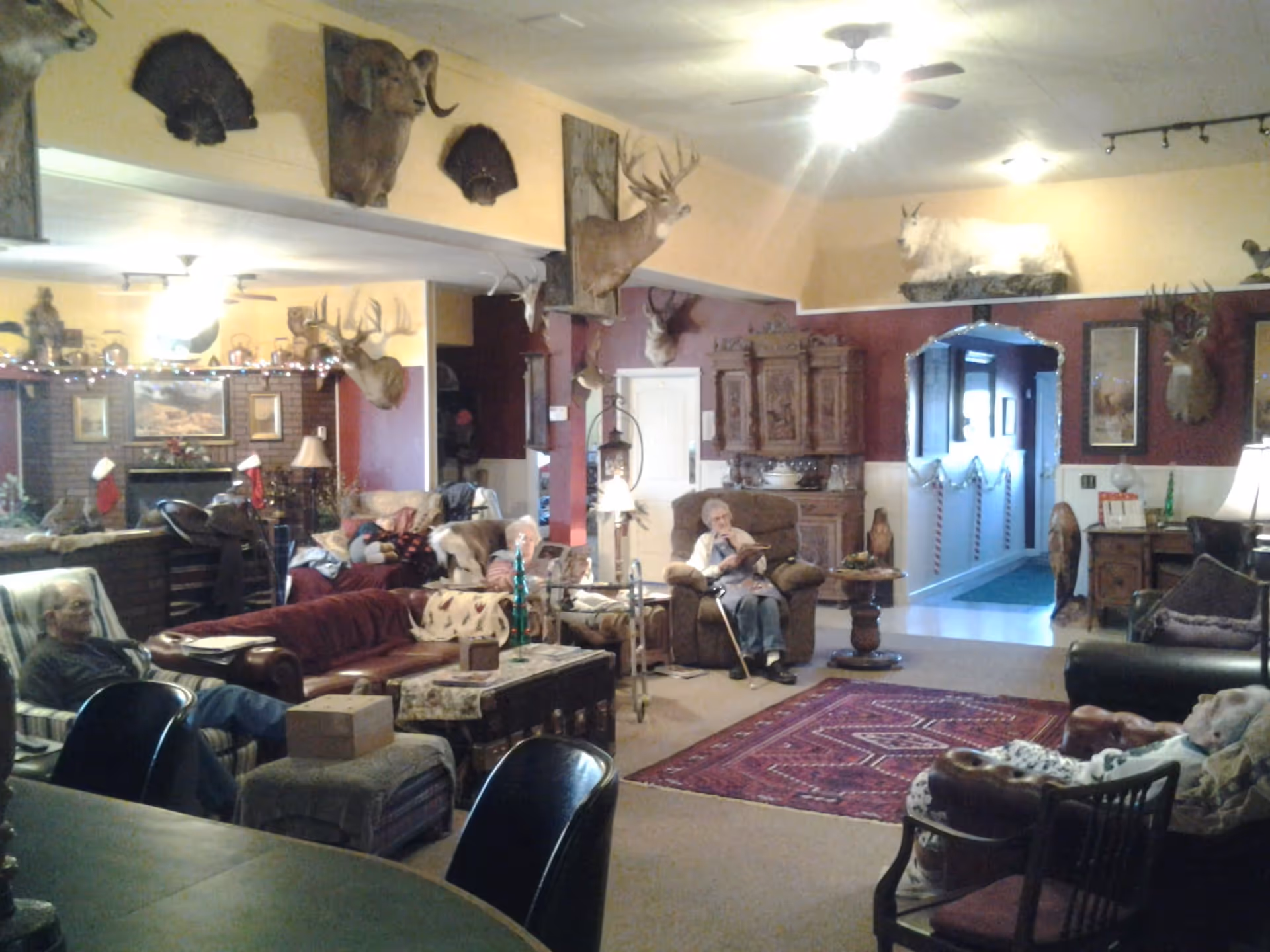 A cozy living room in the Strawberry Roan facility with several elderly residents seated on various chairs and sofas. The room is decorated with numerous mounted animal heads and taxidermy, including deer and a mountain goat. There is a red patterned rug on the floor, wooden furniture, and warm lighting from lamps and ceiling fans.
