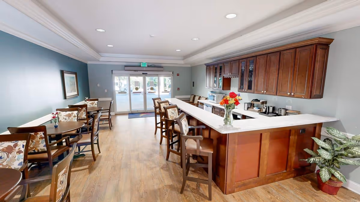 A bright and clean common area with a kitchen and dining space. The kitchen features wooden cabinets, a white countertop with a vase of colorful flowers, and several bar stools. To the left, there are multiple round tables with floral-patterned chairs. The room has light blue walls, wooden flooring, and large glass doors at the far end letting in natural light.